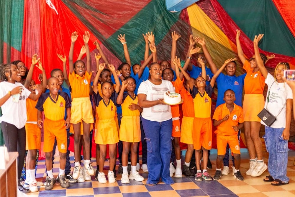 Pupils in orange and blue school uniforms cheering with their arms raised around a cake at a Love & Light Foundation outreach