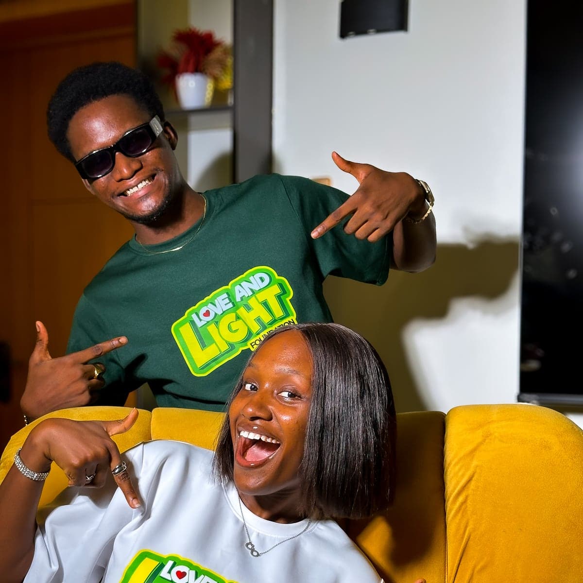 James and Sunmi, two Love & Light Foundation volunteers, smiling and pointing at the foundation logo on their T-shirts